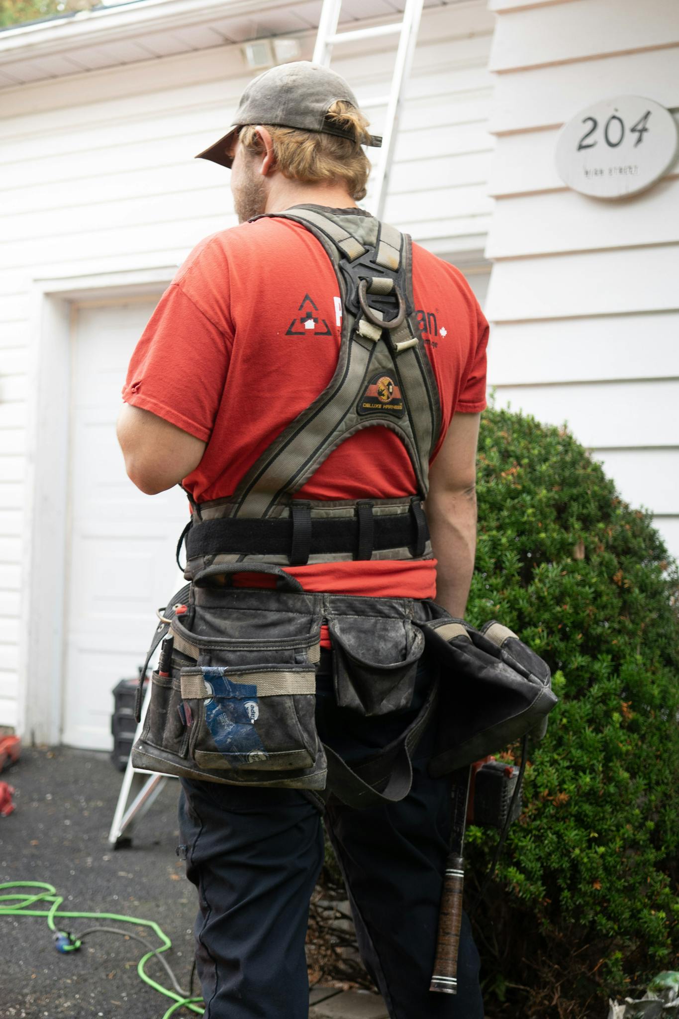 Back view of construction worker in red shirt and safety harness next to a house.
