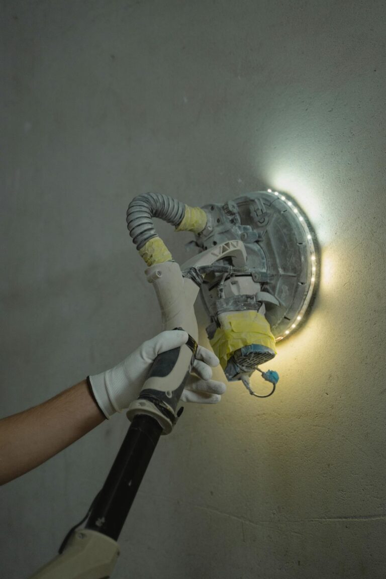 Close-up of a worker sanding a wall with a power tool under bright lighting.