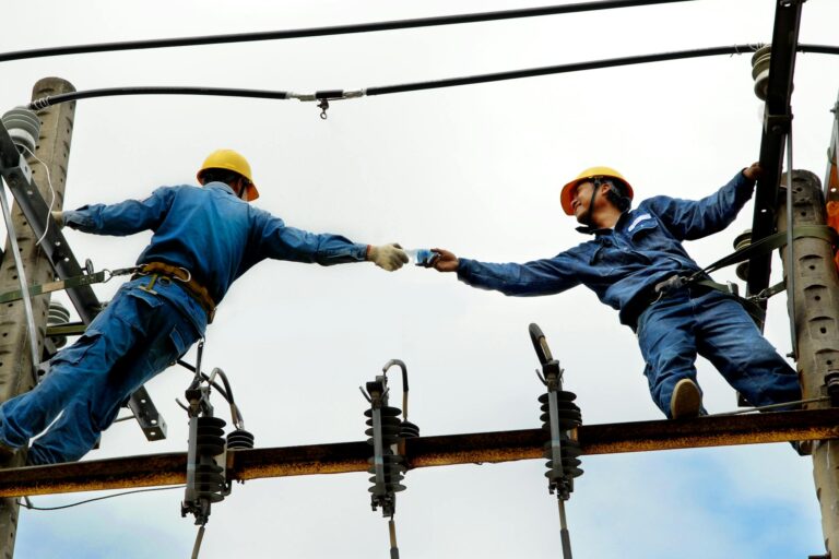 Two electrical workers in blue uniforms collaborating on power lines during daytime.